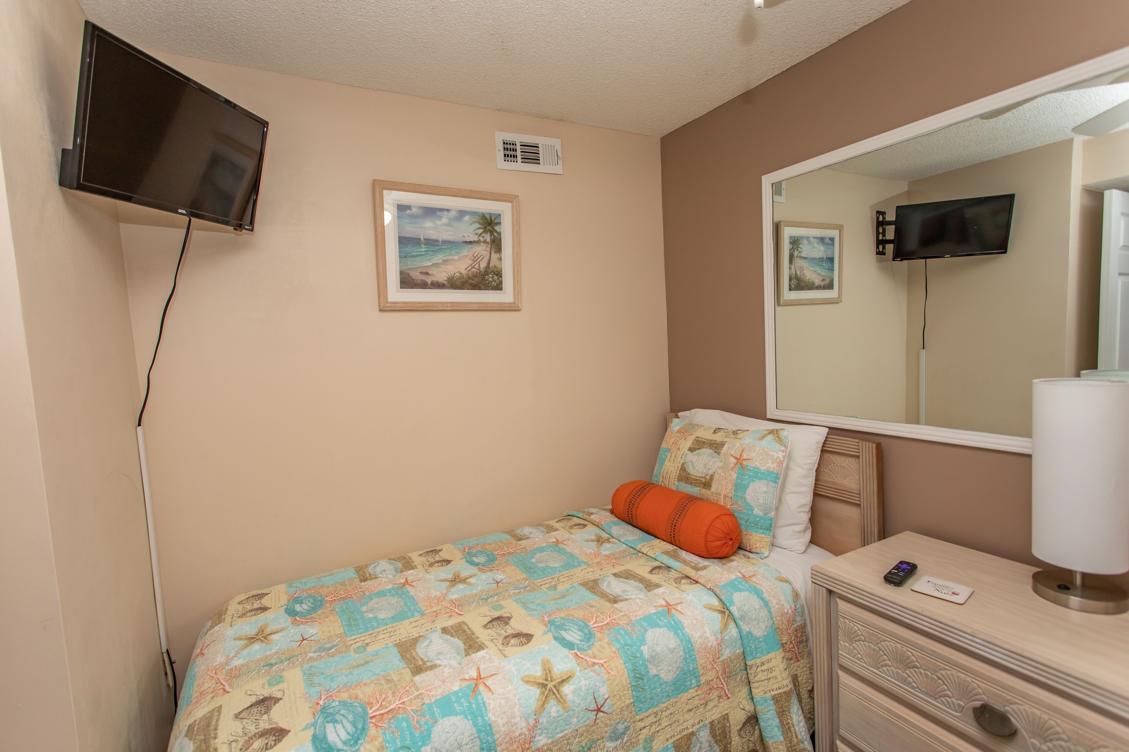 A bedroom with a bed, a bedside table with a lamp, a TV mounted on the wall, and a framed picture of a beach scene.