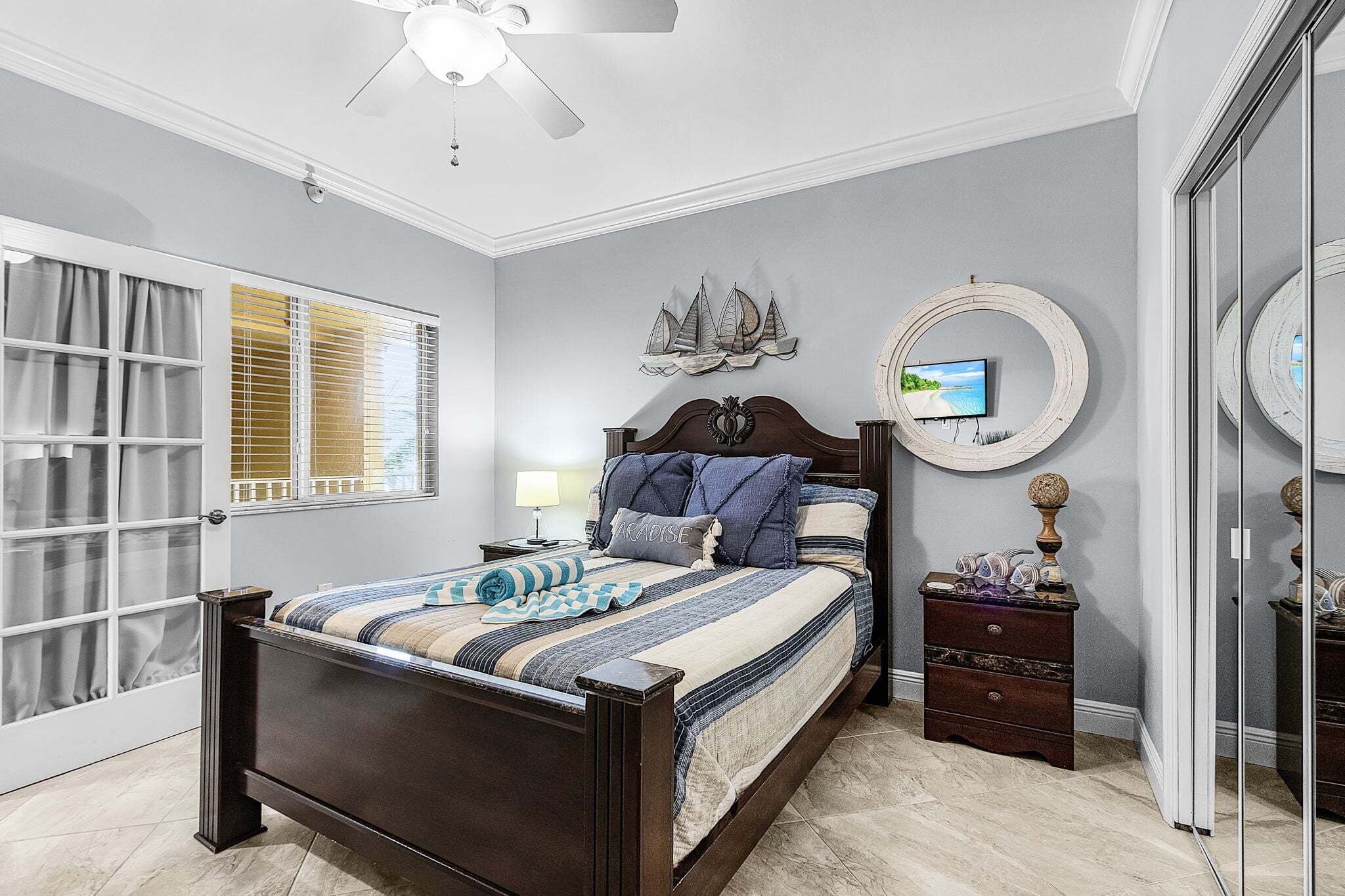 Une chambre à coucher comprenant un lit, une table de chevet, une commode, un ventilateur de plafond et une fenêtre avec des stores.