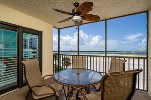 A balcony with a glass table and wicker chairs, overlooking a beach with a clear sky and clouds.
