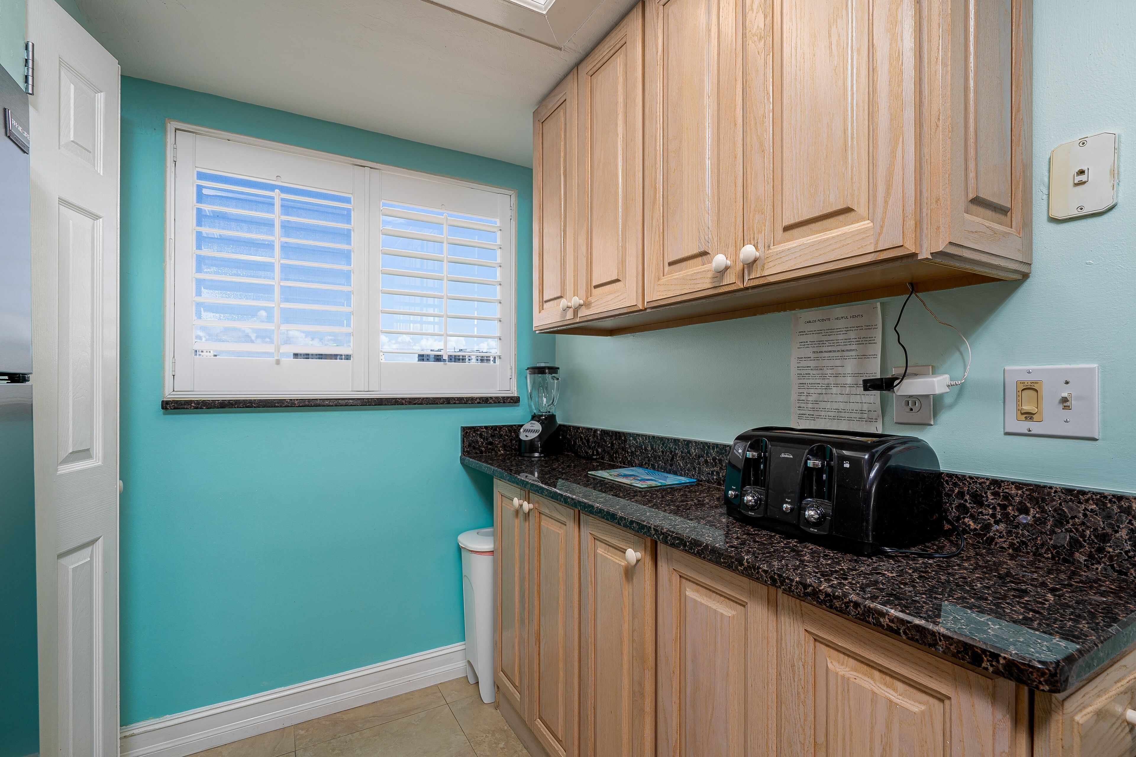 A kitchen with teal walls, wooden cabinets, a granite countertop, a toaster, and a window with blinds.