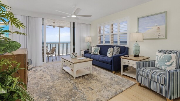 A living room with a blue sofa, a white coffee table, and a striped armchair. There is a balcony with a view of the ocean. - Welcome To Beach Villa's # 406 Vacation Rental - 250 Estero Blvd 2 Bedroom Condo by RedAwning (Fort Myers Beach)