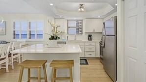 A modern kitchen with white cabinetry, a central island with stools, a stainless steel refrigerator, and a window with blinds. - Welcome To Beach Villa's # 406 Vacation Rental - 250 Estero Blvd 2 Bedroom Condo by RedAwning (Fort Myers Beach)