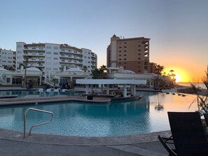 Een zwembadgedeelte van een resort met meerdere zwembaden, ligbedden en een ondergaande zon op de achtergrond.