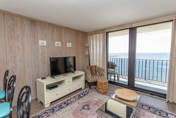 A room with a TV, a sofa, a dining table, and a view of the sea through large windows.
