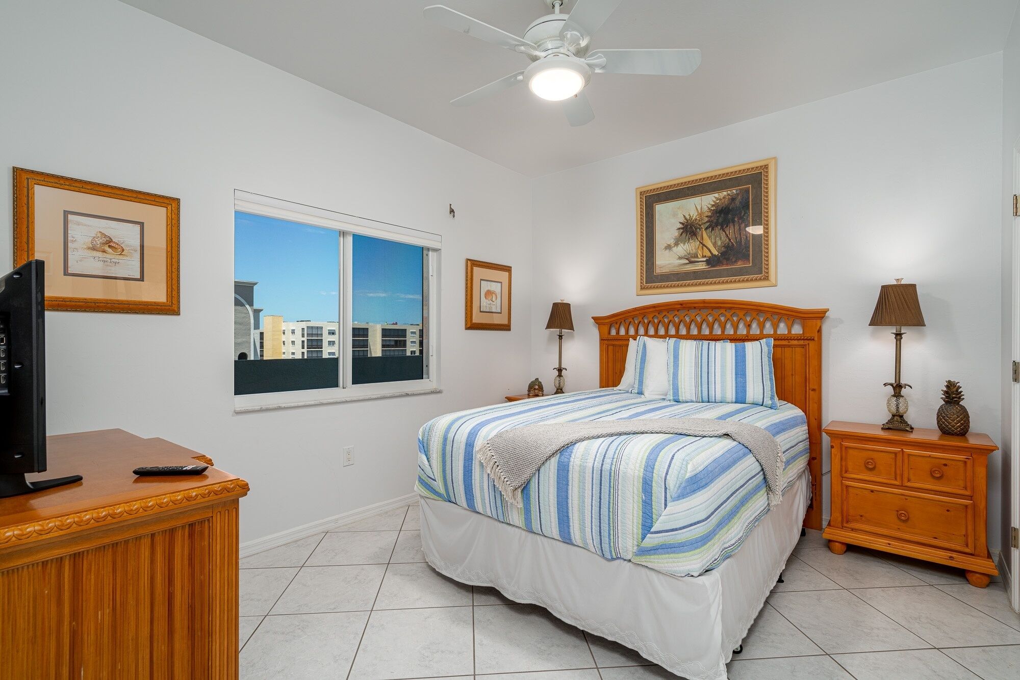 A bedroom with a bed, a television, a window with a city view, a ceiling fan, and framed artwork on the walls.