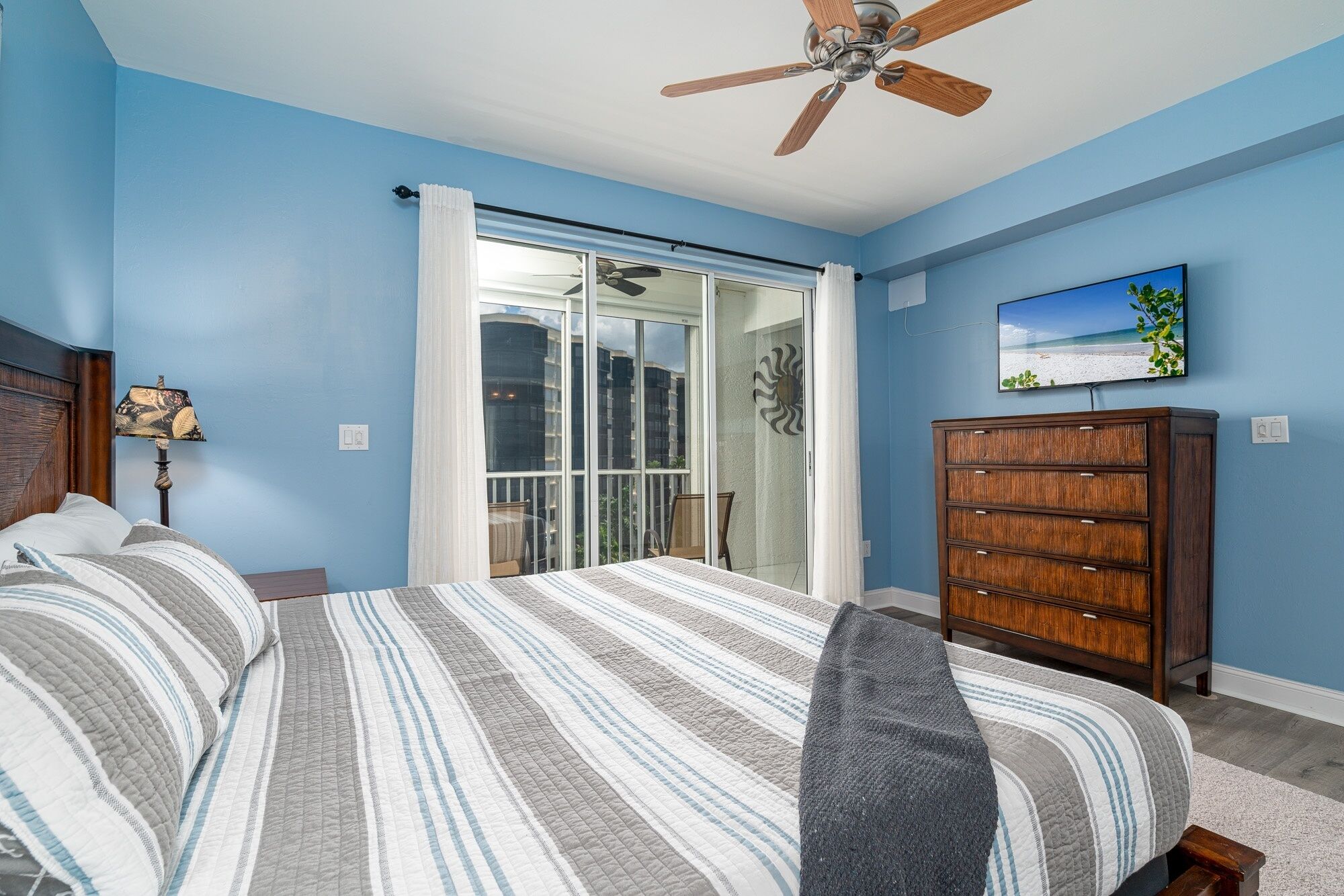 A bedroom with a bed, a dresser, a ceiling fan, a window with curtains, and a painting on the wall.
