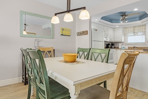Un coin repas avec une table blanche, des chaises vertes et un saladier jaune. On aperçoit, au fond, une cuisine avec des meubles blancs et un plafond bleu.