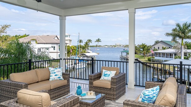 A covered outdoor seating area with wicker furniture and cushions, overlooking a marina with boats and houses. - Magnificent New Build Private Pool Home On The North Tip Of Fort Myers Beach! 5 Bedroom Home by RedAwning (Fort Myers Beach)