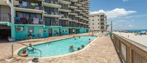 A swimming pool area with a view of a multi-story building, a beach, and a clear sky.