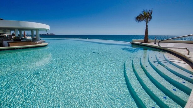 A clear, shallow infinity pool with a palm tree and a white structure with outdoor seating. - Beautiful 2 Bedroom Condo on the Sea of Cortez at Las Palmas Resort D-501 2 Condo by RedAwning (Puerto Peñasco)
