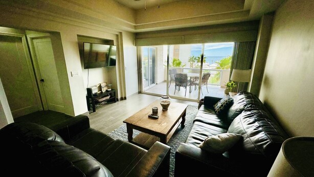 A living room with a leather sofa, a wooden coffee table, and a flat-screen TV. There is a sliding glass door leading to a balcony with outdoor furniture. - Beautiful 1Â½ Bedroom Condo on the Sea of Cortez at Las Palmas Resort D-304 2 Condo by RedAwning (Puerto Peñasco)
