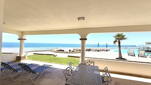 A covered outdoor area with lounge chairs, a pool, and a view of the beach and ocean.