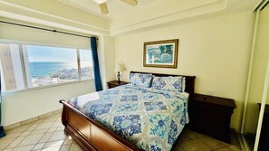 A bedroom with a bed, a nightstand, a lamp, a framed picture, and a window with a view of the sea. - Spectacular 2 Bedroom Condo on Sandy Beach at Las Palmas Resort D-505 2 Condo by RedAwning (Puerto Peñasco)
