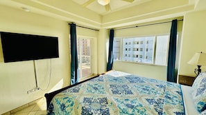 Habitación de hotel con cama, televisor montado en la pared, ventana con cortinas y vista a edificios.