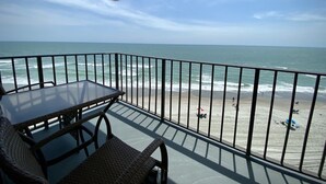 Ein Balkon mit Tisch und Stühlen mit Blick auf einen Strand, auf dem sich Menschen und Sonnenschirme befinden.