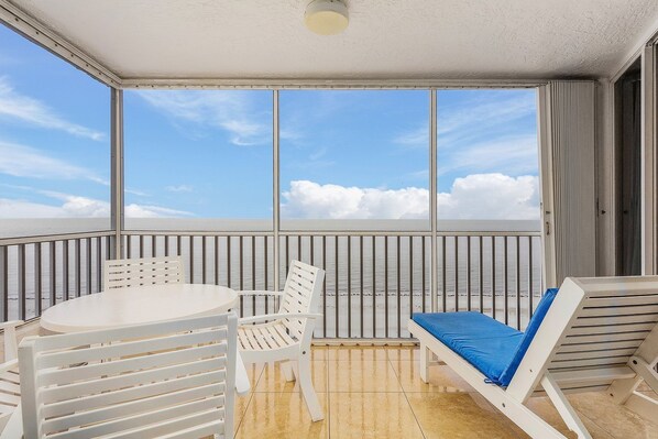 A balcony with a white table and chairs, a blue cushioned lounge chair, and a view of the sea. - Beautiful Penthouse Condo - Corner Unit! 2 Bedroom Condo by RedAwning (Fort Myers Beach)