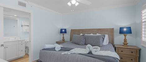 Une chambre à coucher comprenant un lit, deux tables de chevet avec des lampes, un ventilateur de plafond, une tête de lit en bois, et une salle de bain avec un miroir et des placards.