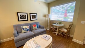 A living room with a grey sofa, two framed pictures on the wall, a small round table with a lamp, and a window with blinds. - Harbour House At The Inn Unit 211 1 Bedroom Condo by RedAwning (Fort Myers Beach)