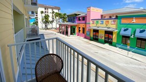 A balcony with a wicker chair, a street with colorful buildings, and palm trees. - Harbour House At The Inn Unit 211 1 Bedroom Condo by RedAwning (Fort Myers Beach)