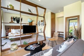 A modern living room with a sofa, a coffee table, and a TV. There is a bookshelf with decorative items and a bed with a headboard in the background.