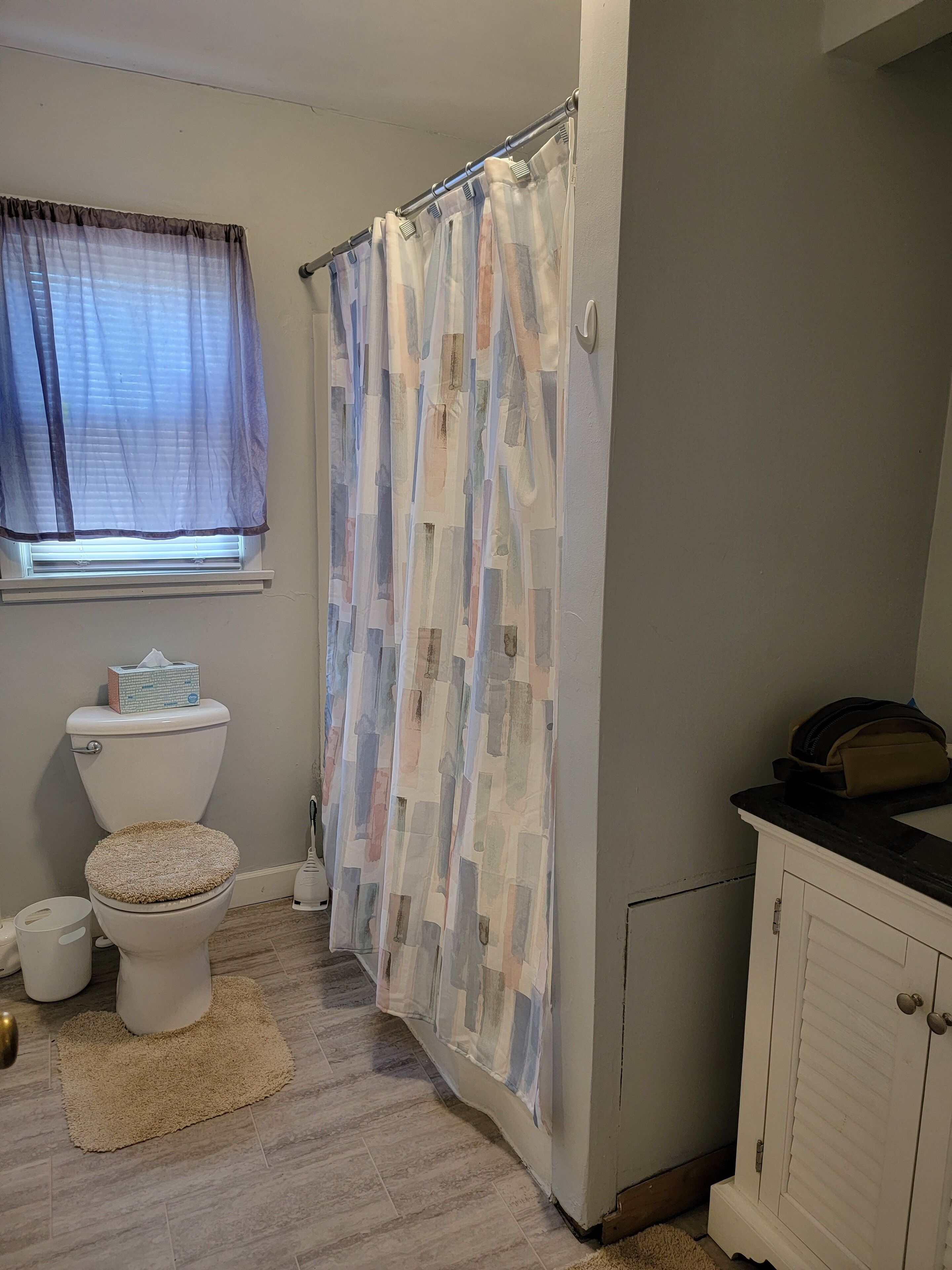 Een badkamer met een toilet, douchegordijn, raam met zonwering en een kast.