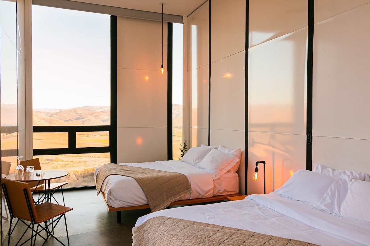 Een hotelkamer met twee bedden, een zithoek met een tafel en stoelen, en een groot raam met uitzicht op het landschap.