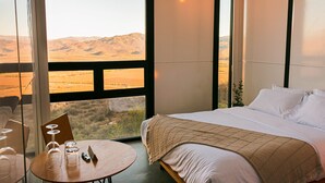 Een slaapkamer met een groot raam, een bed, een rond tafeltje met glazen en uitzicht op een woestijnlandschap.