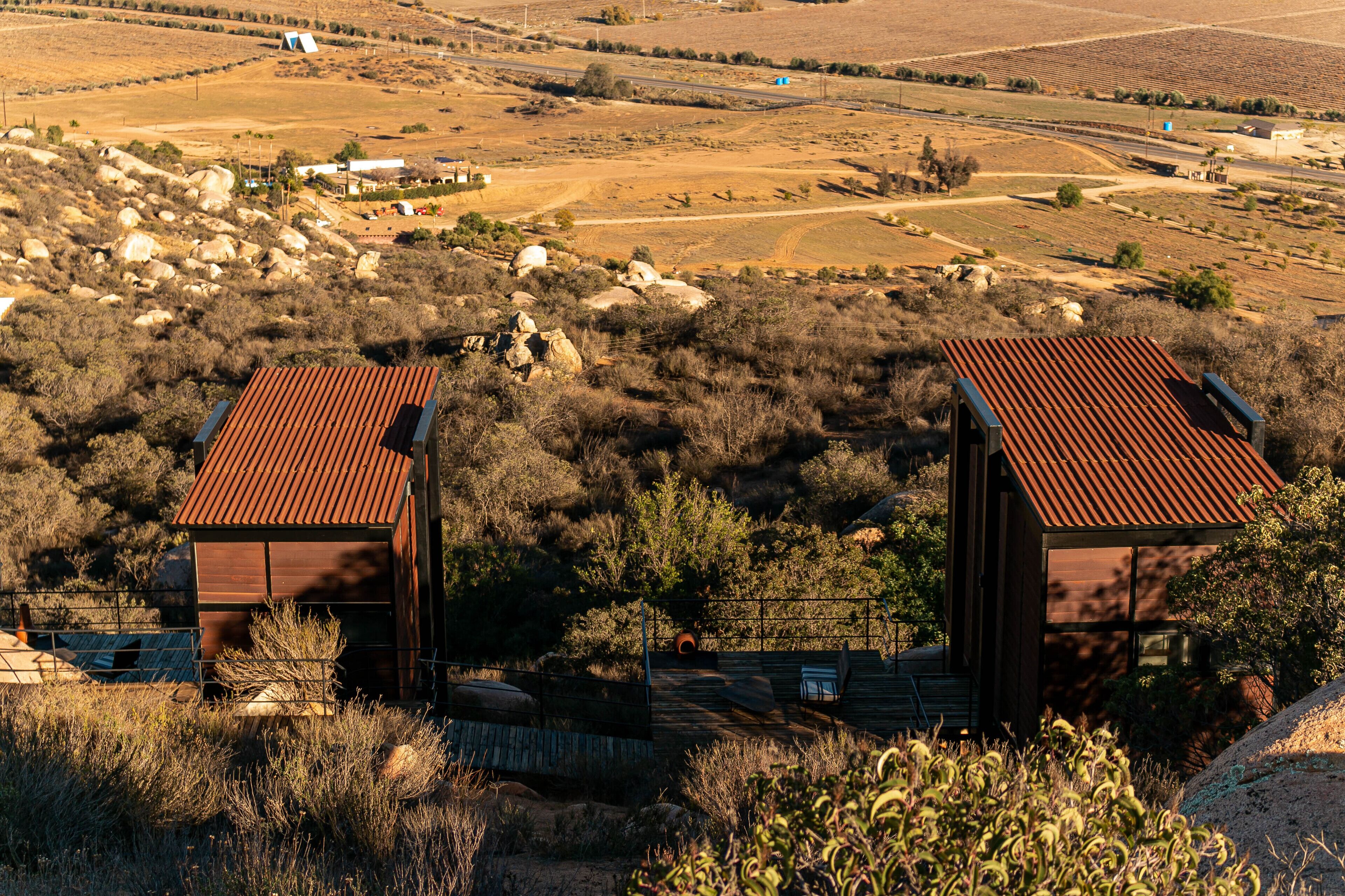 Deux cabanes rustiques aux toits métalliques, entourées d’une végétation aride et d’un terrain rocheux, et offrant une vue imprenable sur un vaste paysage.