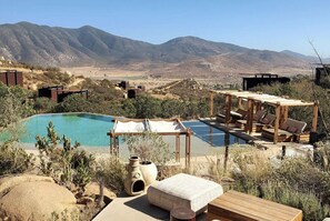 Une piscine dotée d’une terrasse en bois et de chaises longues, entourée d’une végétation désertique et avec des montagnes en arrière-plan.