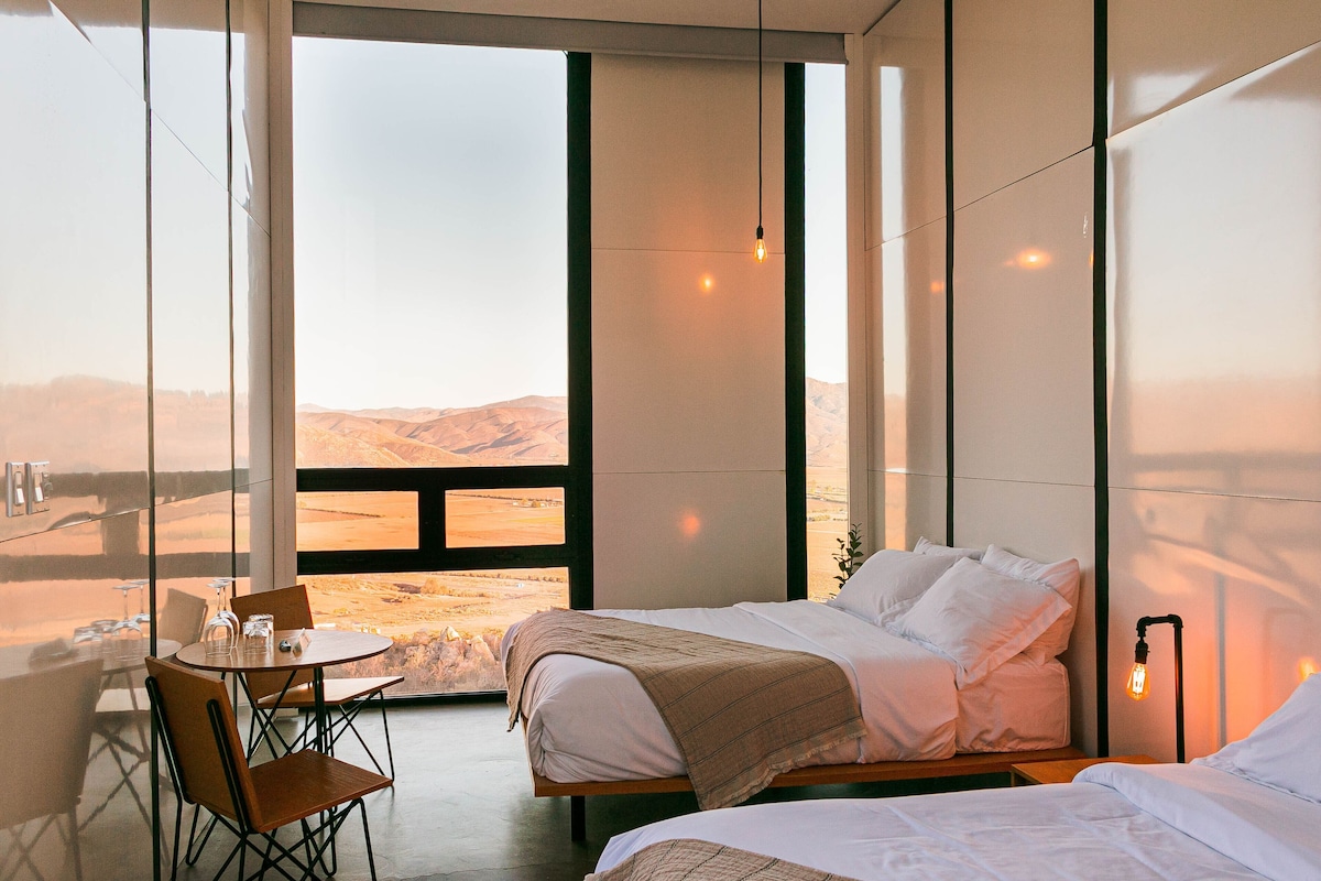 Een moderne hotelkamer met een groot raam, een bed, een nachtkastje met glazen en een stoel.