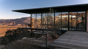 Een modern huis met een houten terras, grote glasramen en uitzicht op een bergachtig landschap.