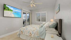 A bedroom with a bed, a ceiling fan, a TV, a window with blinds, and a balcony door.