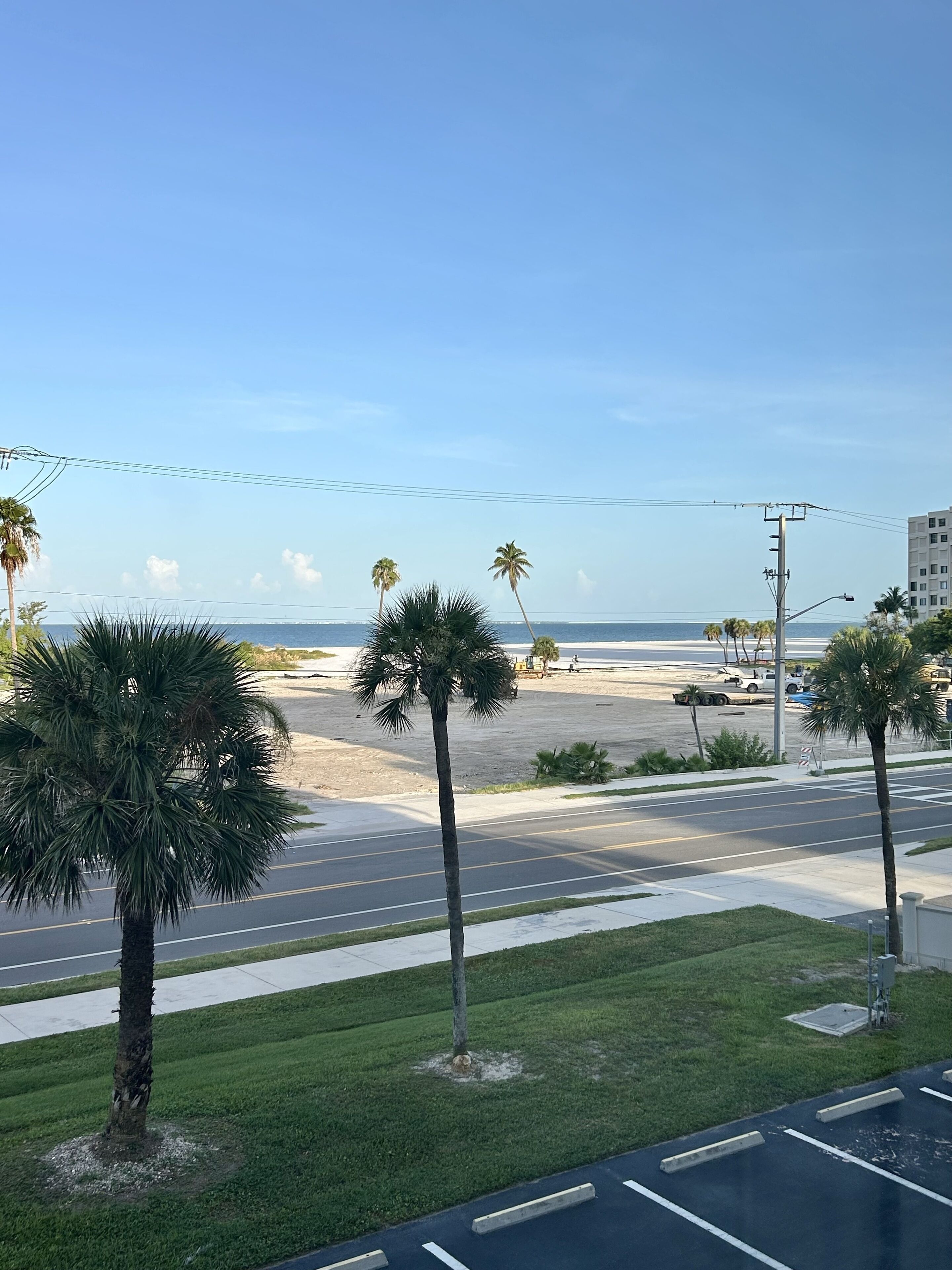 A street view with palm trees, a beach, and a clear sky.