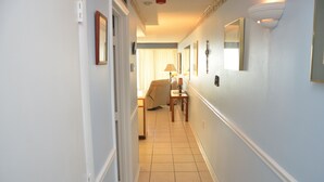 A narrow hallway with a chair and a table, a door on the left, and a wall with a mirror and a light fixture. - Royal Garden Resort 1401 3 Bedroom Condo by RedAwning (Murrells Inlet)