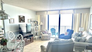 A spacious living room with a television, white furniture, a balcony with chairs, and a large window with curtains. - Royal Garden Resort 1401 3 Bedroom Condo by RedAwning (Murrells Inlet)