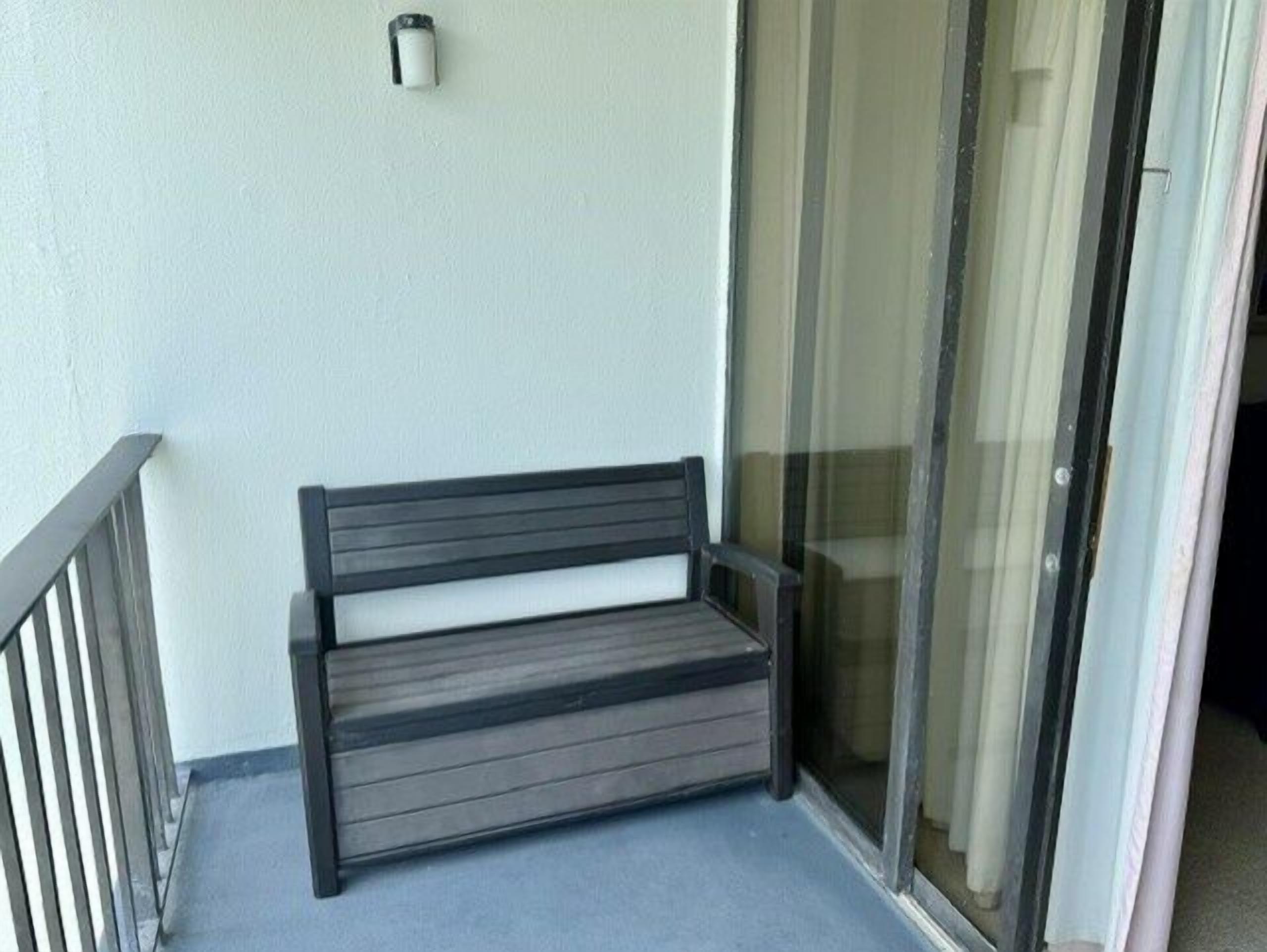 Een bankje op een balkon, met een glazen deur en een klein, aan de muur bevestigd lampje.