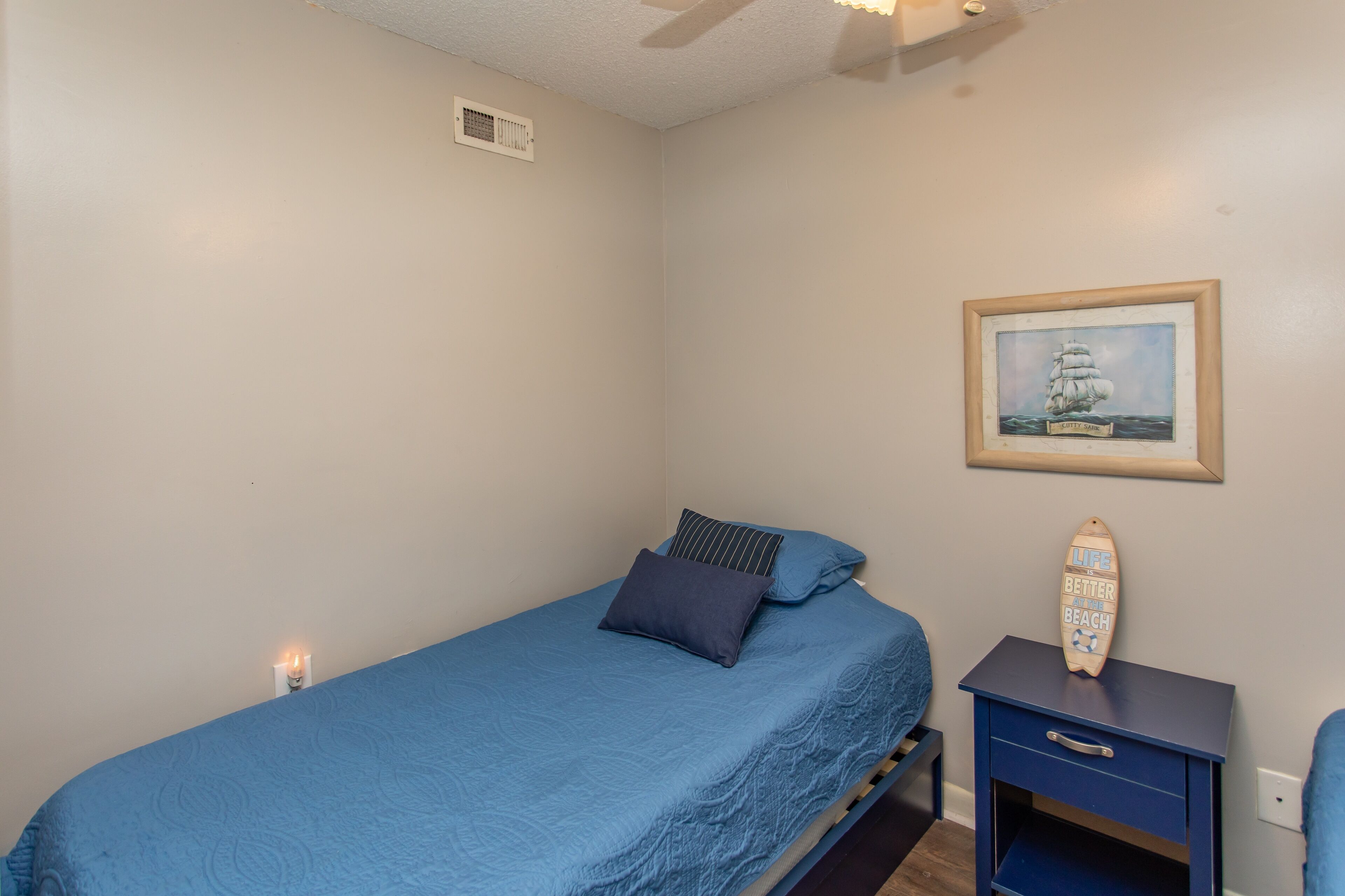 A bedroom with a single bed, a nightstand with a drawer, a framed picture on the wall, and a small shelf with a decorative item.