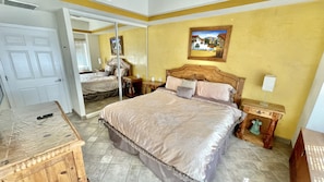 A bedroom with a large bed, a wooden nightstand, a bedside table, a mirror, and a door leading to another room. - Beautiful 1Â½ Bedroom Condo on the Sea of Cortez at Las Palmas Resort D-302 2 Condo by RedAwning (Puerto Peñasco)