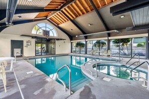 Una piscina interior con paredes de vidrio transparente, techo de madera y zona de estar con mesas y sillas.