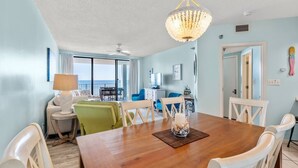 A dining area with a wooden table, white chairs, and a chandelier. There is a window with a view of the outside. - Pelican Pointe|Beachfront|Gulf Views|Indoor Pool (Orange Beach)