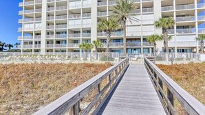 A multi-story hotel with balconies, a wooden boardwalk leading to the entrance, and palm trees. - Pelican Pointe|Beachfront|Gulf Views|Indoor Pool (Orange Beach)
