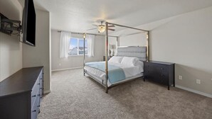 A bedroom with a four-poster bed, a ceiling fan, a TV mounted on the wall, and a window with curtains. - Modern Family Friendly House (Kennewick)