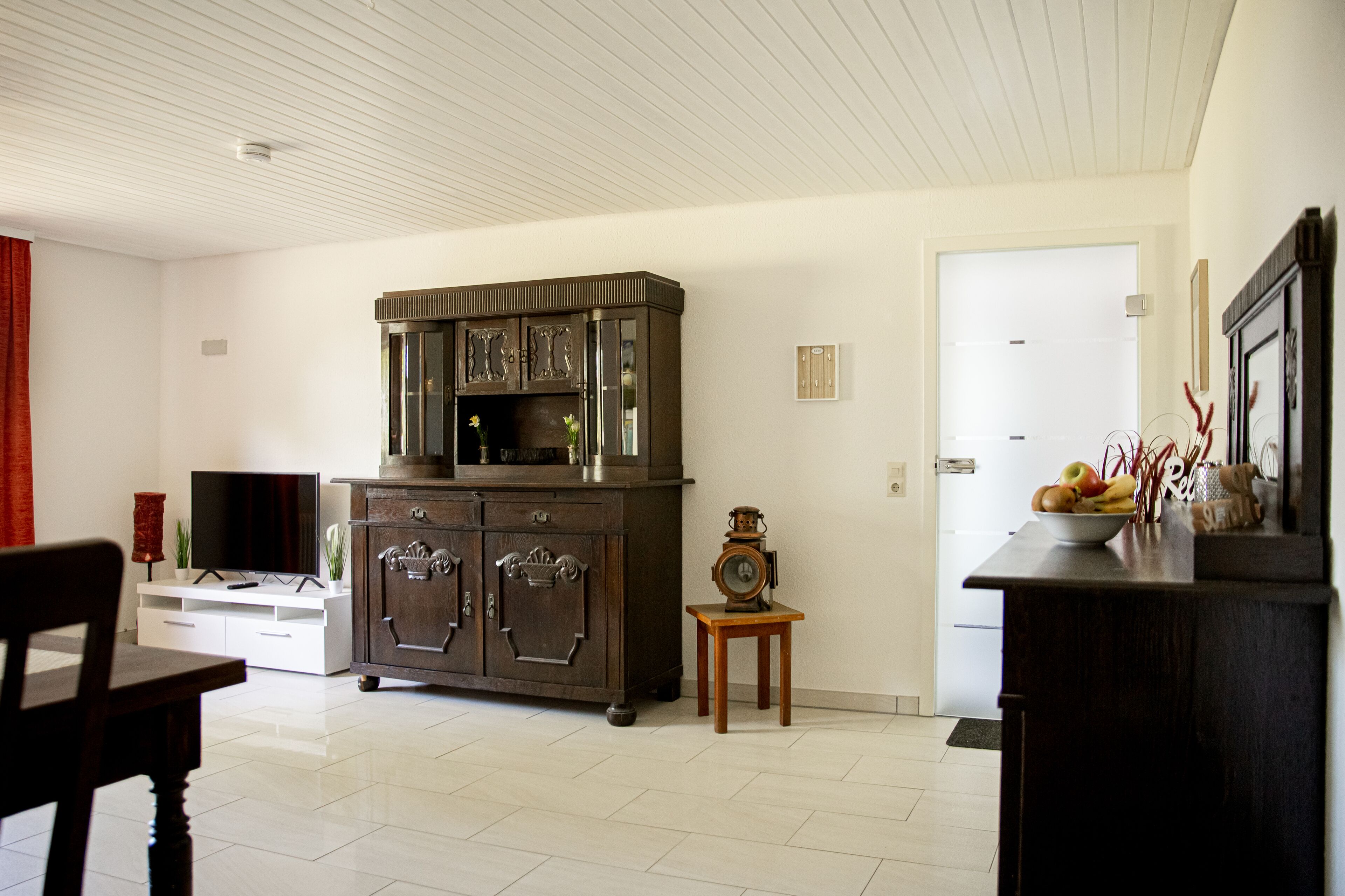 Un salon comprenant un meuble en bois foncé, une petite table en bois avec une horloge, et un meuble de rangement blanc avec un téléviseur à écran plat.