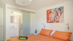 Ein modernes Hotelzimmer mit einem großen Bett, orangefarbener Bettwäsche, einem Bild an der Wand und einem Badezimmer mit Spiegel und Waschbecken.