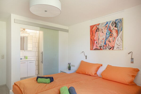 A modern hotel room with a large bed, orange bedding, a painting on the wall, and a bathroom with a mirror and sink.