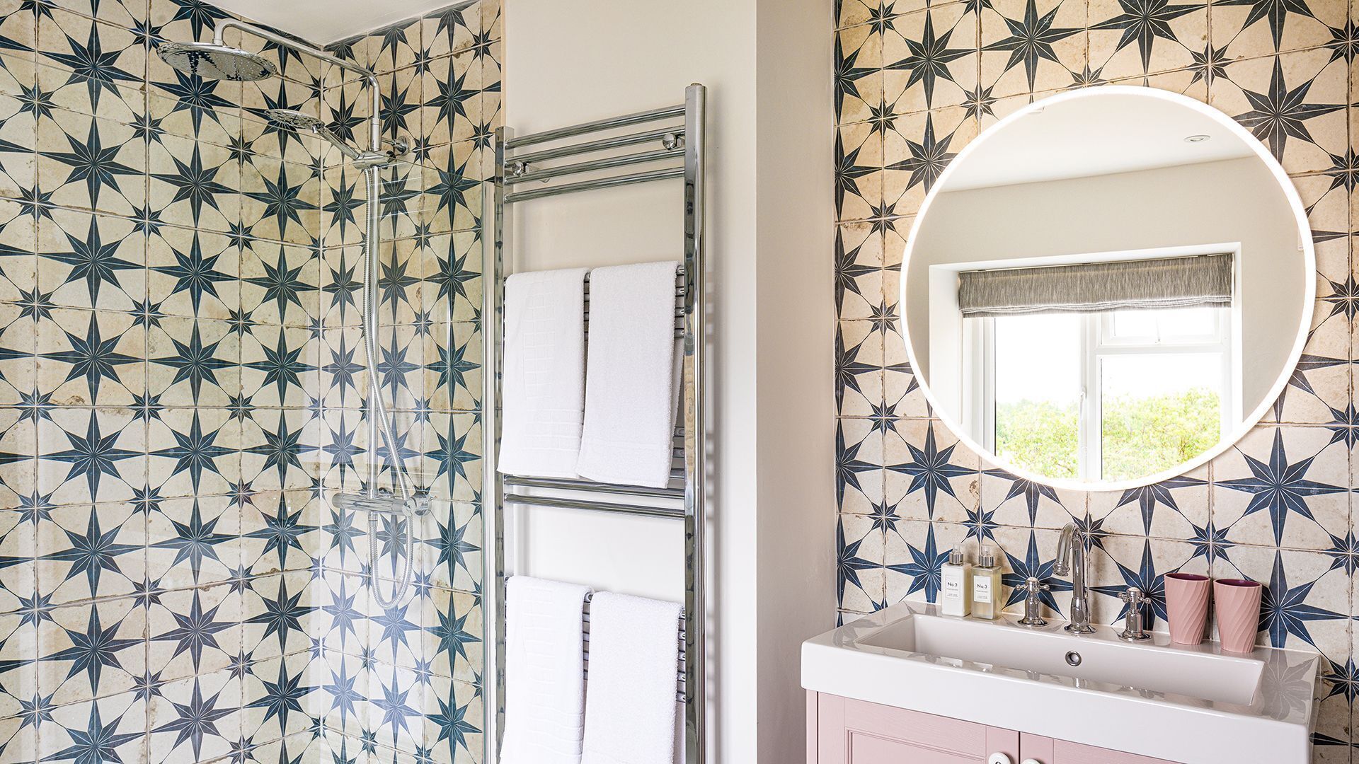A bathroom with star-patterned tiles, a shower with a glass door, a round mirror, and a window with a view of greenery.