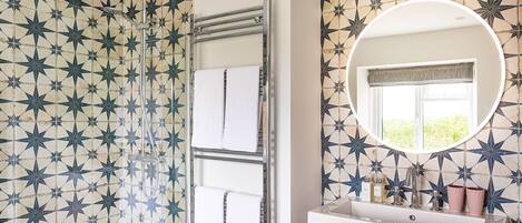 A bathroom with star-patterned tiles, a shower with a glass door, a round mirror, and a window with a view of greenery.