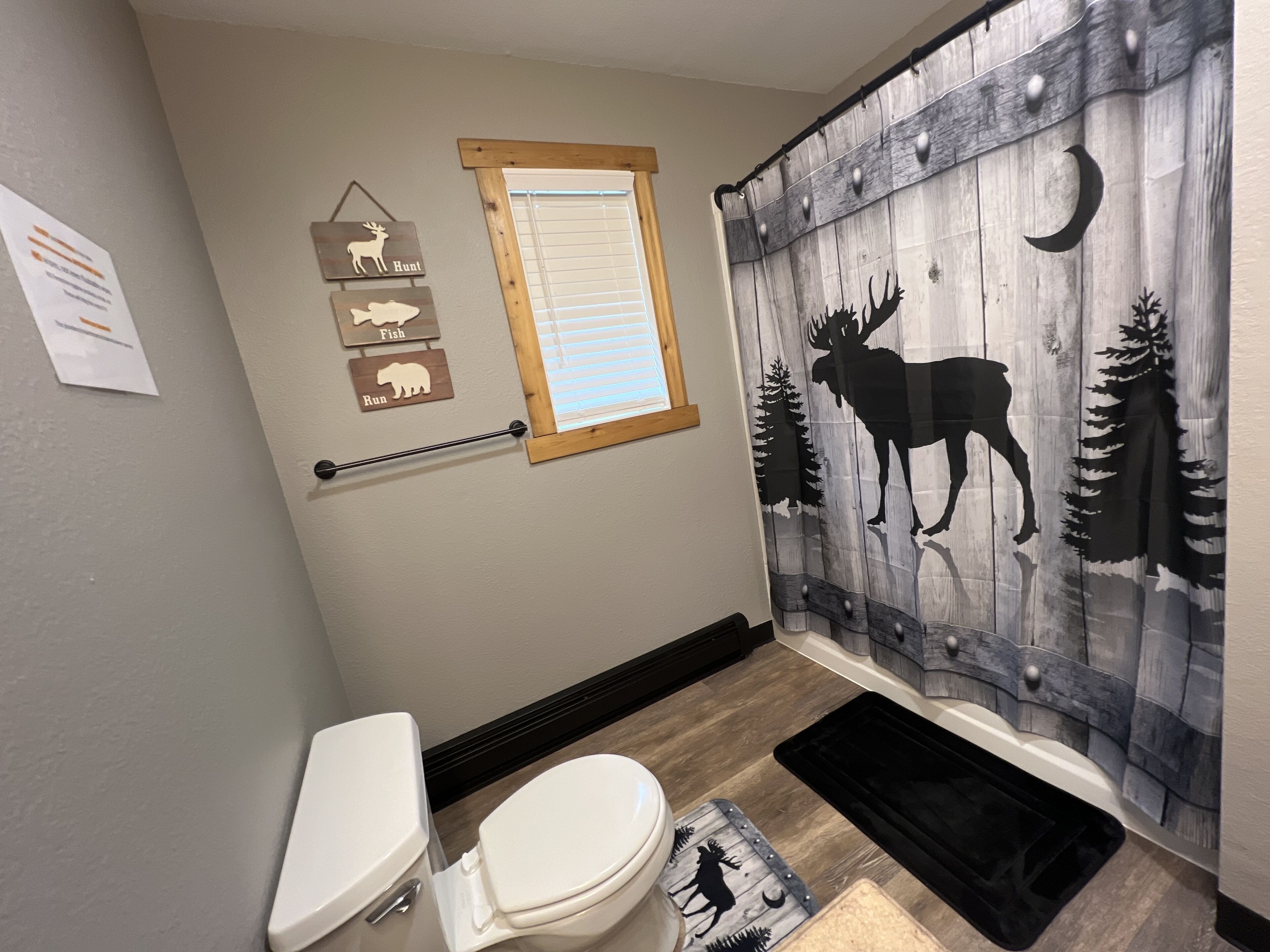 A bathroom with a toilet, a window, a shower curtain with a forest and moose design, and a wall-mounted wooden sign with animal silhouettes.