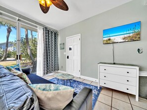 A room with a bed, a white dresser, a painting of a beach, and a view of palm trees through the window.