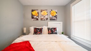 Ein ordentlich bezogenes Bett mit weißer Bettwäsche und einer roten Tagesdecke, ein Nachttisch mit einer Lampe sowie ein Fenster mit Jalousien.